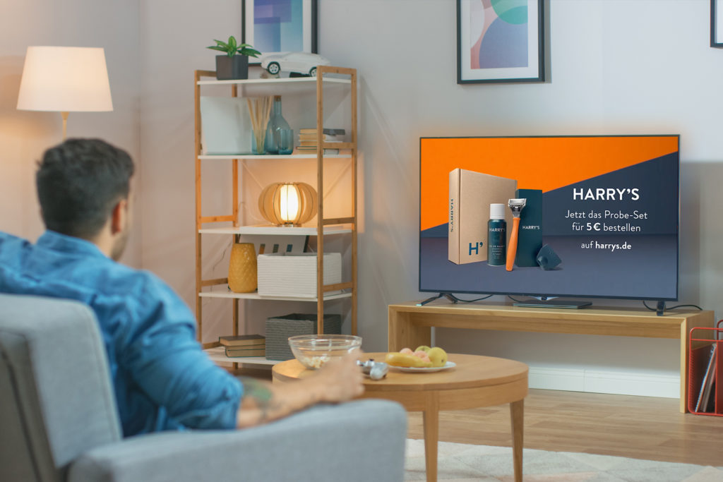 Ein Mann sitzt auf einem Sofa in einem gemütlich eingerichteten Wohnzimmer und sieht einen Werbespot für das Harry's Rasierset im Fernsehen.