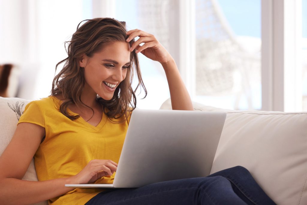 Frau mit gelbem Oberteil sitzt entspannt auf einem Sofa und arbeitet lächelnd an einem Laptop.