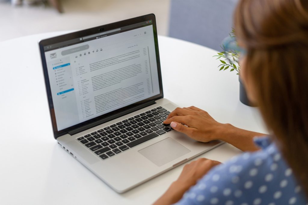 Eine Person sitzt an einem weißen Tisch und arbeitet an einem Laptop, auf dessen Bildschirm eine E-Mail-Anwendung geöffnet ist.