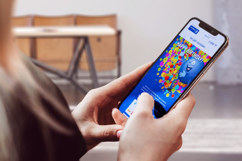 Eine Person hält ein Smartphone, auf dem eine bunte Geburtstagsnachricht angezeigt wird. Der Bildschirm zeigt einen farbenfrohen Hintergrund mit Luftballons und einem animierten Charakter sowie den Text "Happy Birthday!".
