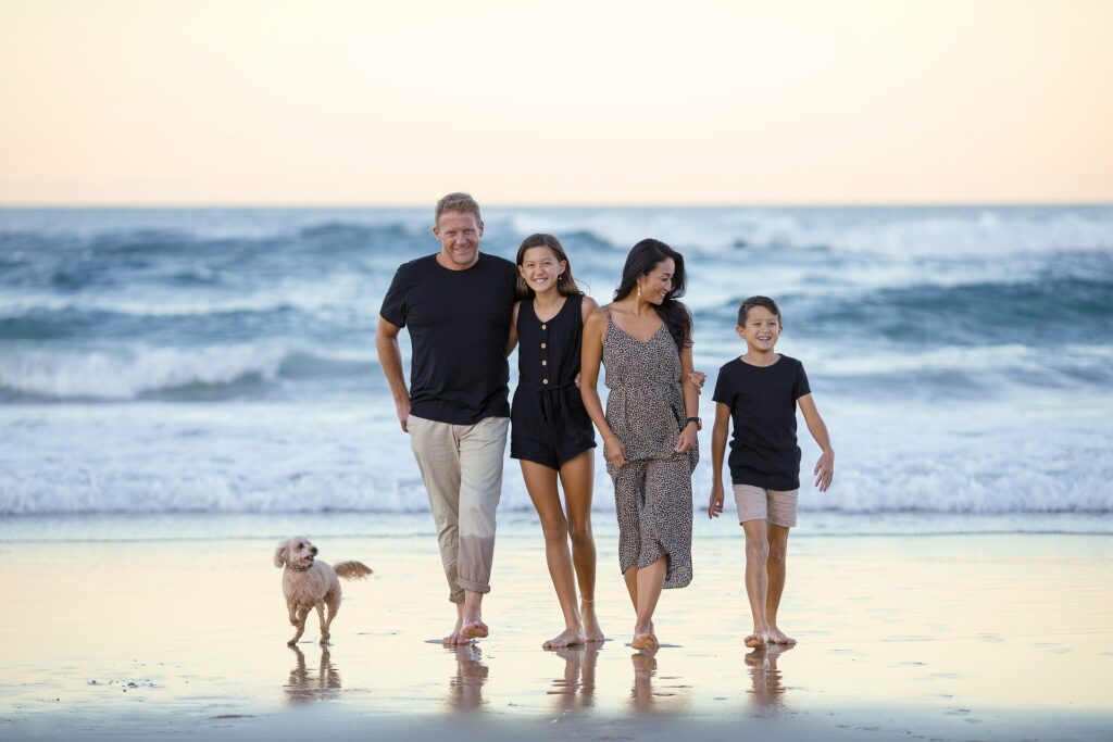 Eine vierköpfige Familie geht lächelnd barfuß am Strand entlang, begleitet von einem kleinen Hund. Im Hintergrund sind Wellen und ein sanfter, heller Himmel zu sehen. Die Szene vermittelt ein Gefühl von Harmonie, Entspannung und Familienzeit in einer natürlichen Umgebung.