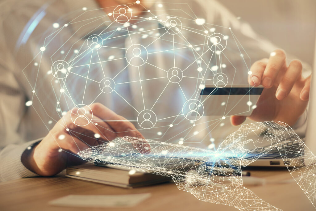 Eine Person hält eine Kreditkarte und tippt auf einem Tablet, während eine digitale Illustration darübergelegt ist. Die Illustration zeigt ein Netzwerk aus verbundenen Punkten und Symbolen von Personen, die eine globale digitale Vernetzung darstellen. Im Hintergrund ist ein Laptop sichtbar. Das Bild symbolisiert Online-Transaktionen und digitale Netzwerke.
