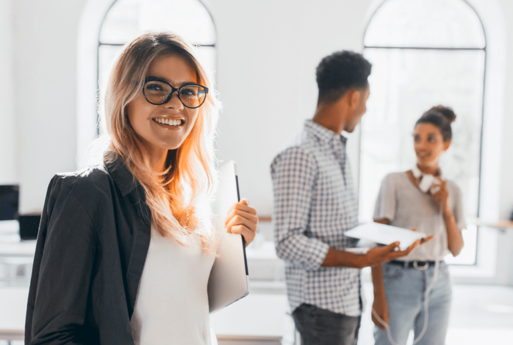 Eine lächelnde Frau mit Brille und blonden Haaren steht im Vordergrund, hält ein Notizbuch und schaut zur Kamera. Im Hintergrund unterhalten sich zwei weitere Personen in einem modernen Büro.