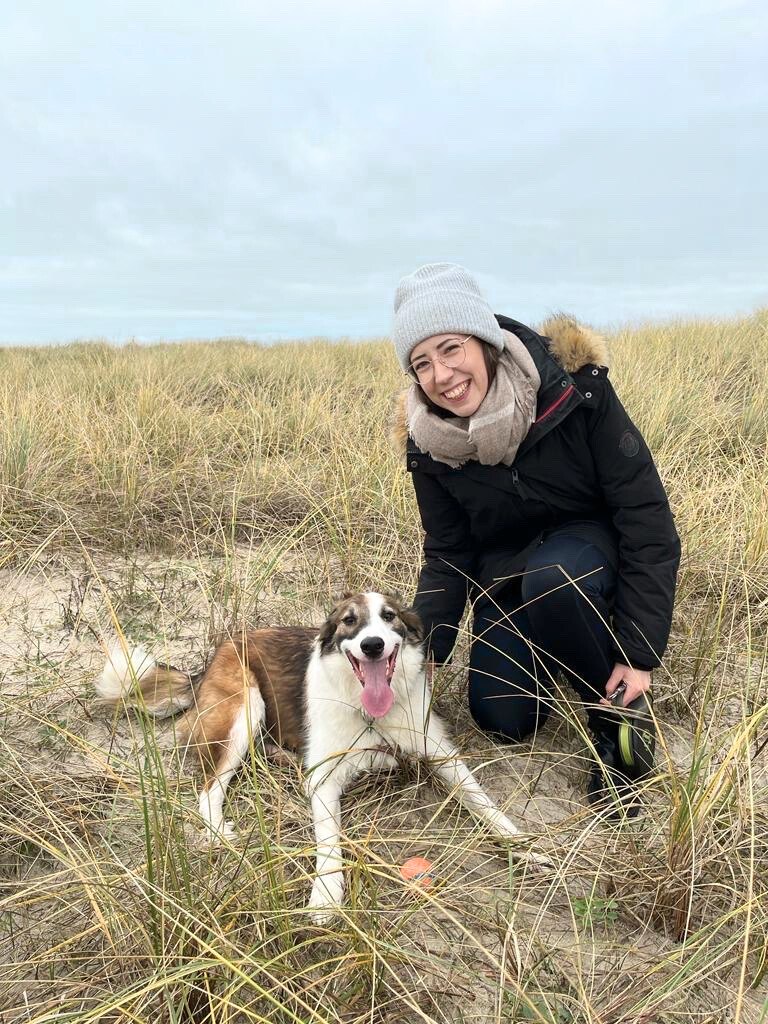 Celina Behrens mit Brille, grauer Mütze und schwarzer Winterjacke kniet in den Dünen und lächelt in die Kamera, während ein großer, fröhlicher Hund mit braun-weißem Fell und herausgestreckter Zunge davor liegt.