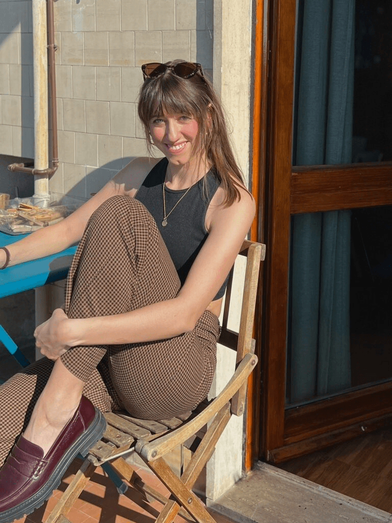 Angelina Michna mit langen braunen Haaren sitzt lächelnd auf einem Holzstuhl auf einer sonnigen Terrasse. Sie trägt ein schwarzes Oberteil, karierte braune Hosen und weinrote Loafer. Eine Sonnenbrille ist auf ihrem Kopf platziert.