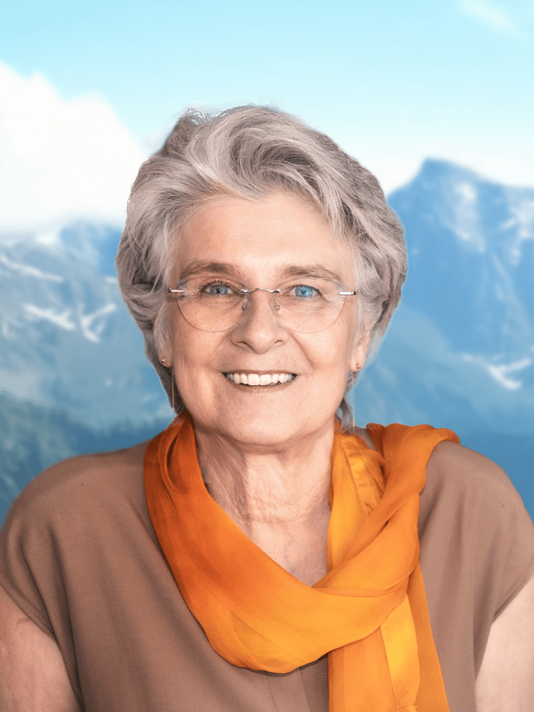 Barbara Wüstenberg mit grauem Haar und Brille lächelt in die Kamera. Sie trägt einen orangefarbenen Schal und ein beigefarbenes Oberteil. Im Hintergrund sind unscharf verschneite Berggipfel und ein blauer Himmel zu sehen.
