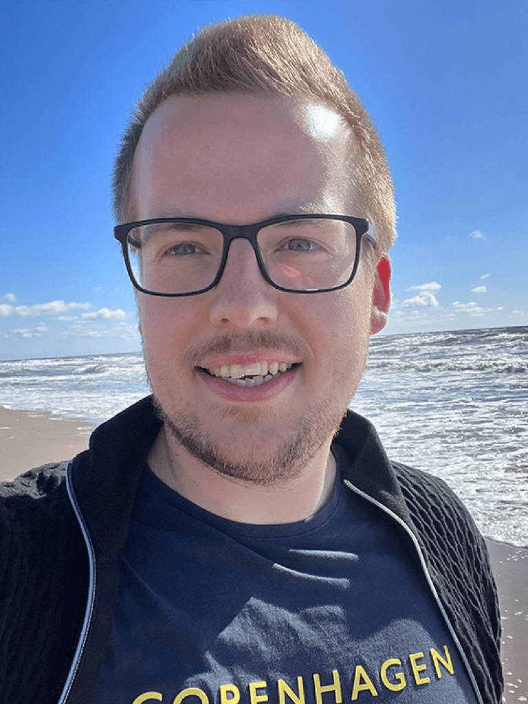 Björn Michelsen mit blondem Haar, Brille und einem blauen T-Shirt steht lächelnd am Strand, während die Sonne scheint. Im Hintergrund sind Meer und blauer Himmel zu sehen.