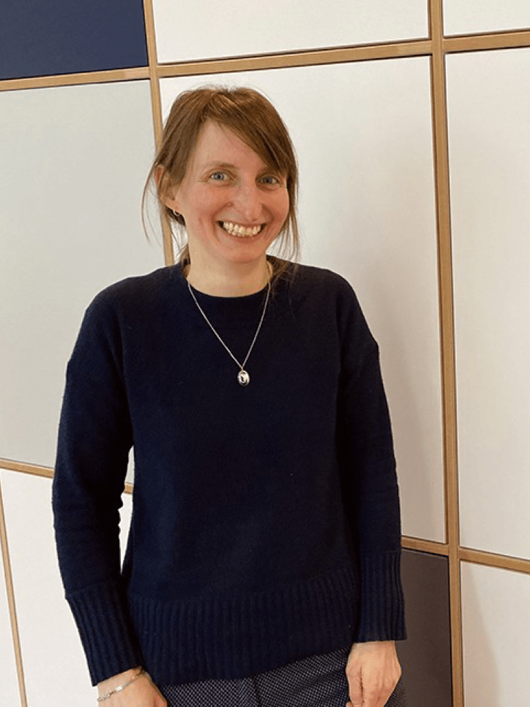 Claudia Sander mit braunen Haaren und einem dunkelblauen Pullover steht vor einer hellen, modernen Wand mit Holzrahmen. Sie lächelt freundlich in die Kamera und trägt eine Silberkette.