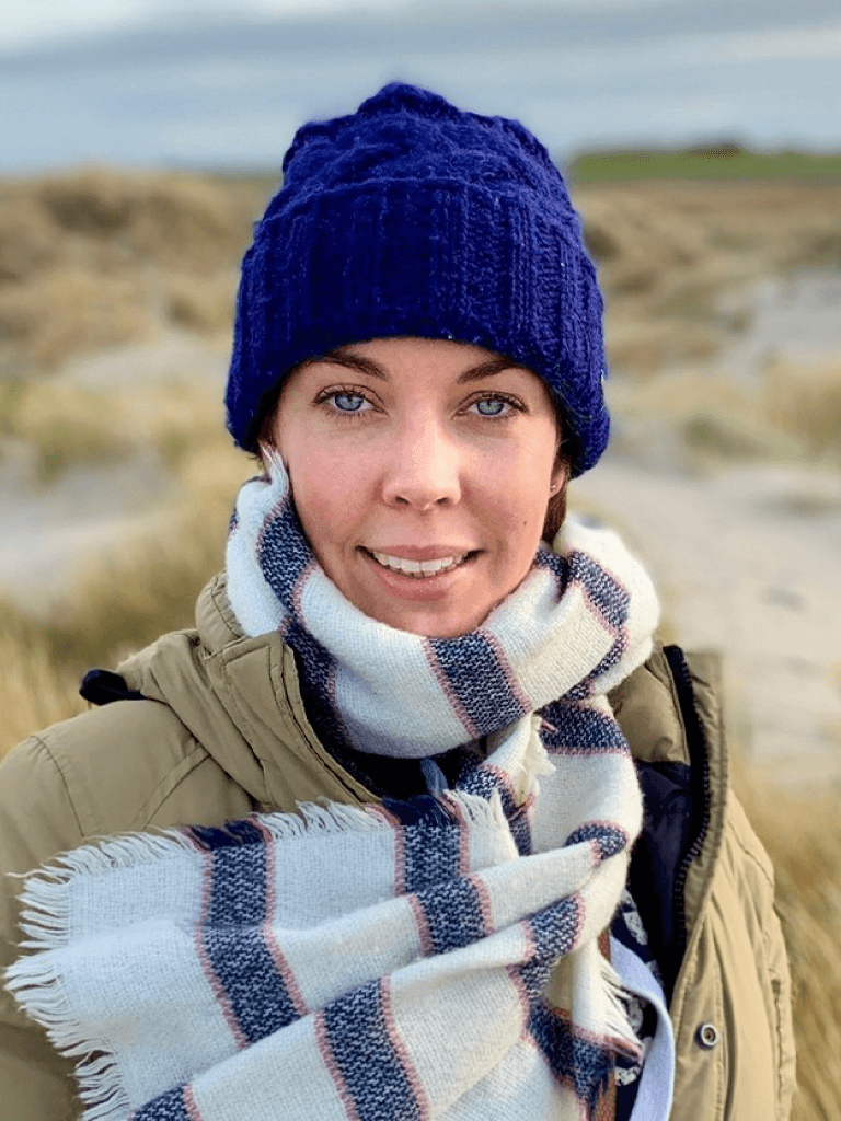 Hilke Götz mit blauer Strickmütze und Schal steht in einer Dünenlandschaft. Sie trägt eine beige Jacke und lächelt in die Kamera.