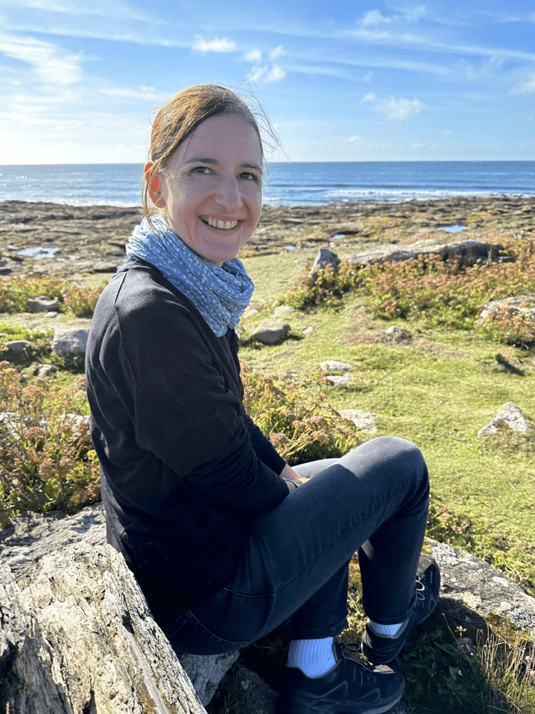 Katja Fangrad mit einem blauen Schal und dunkler Kleidung sitzt auf einem Felsen in einer Küstenlandschaft. Sie lächelt in die Kamera, während im Hintergrund das Meer, eine felsige Küste und ein blauer Himmel mit wenigen Wolken zu sehen sind.