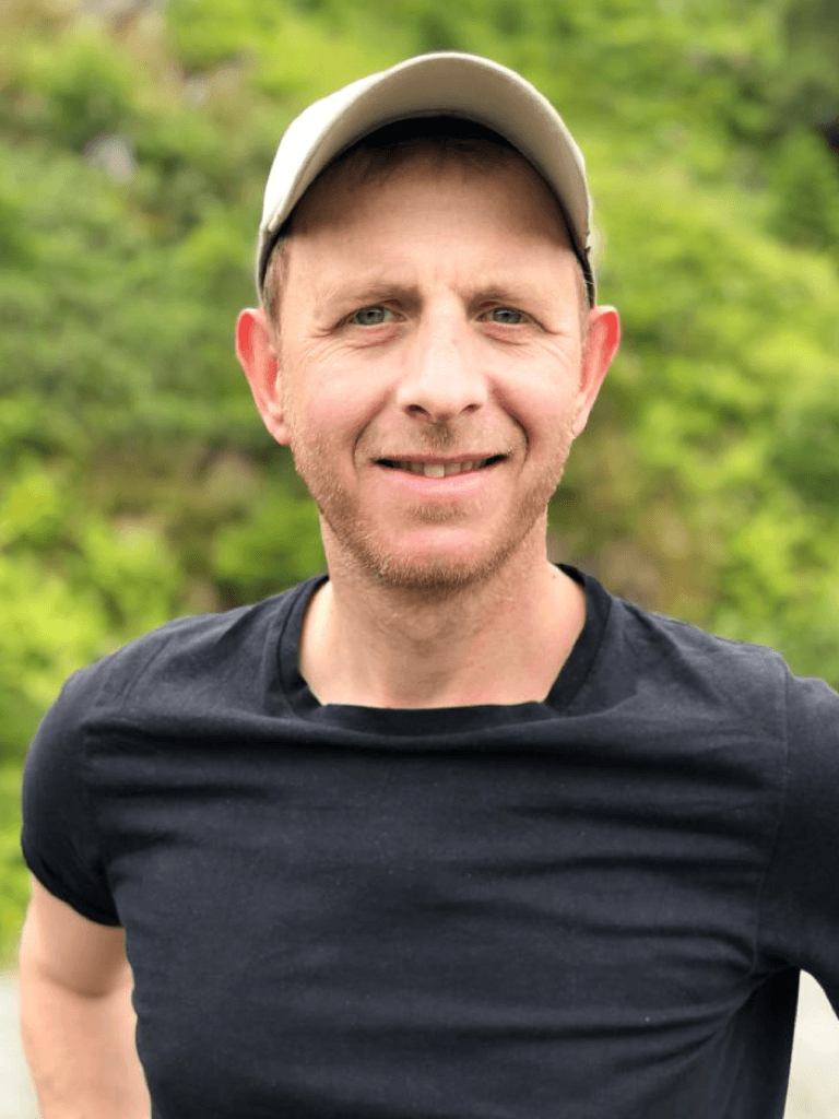 Klaus Poerschke mit weißer Baseballmütze und schwarzem T-Shirt steht vor einer grünen Naturkulisse und lächelt freundlich in die Kamera.