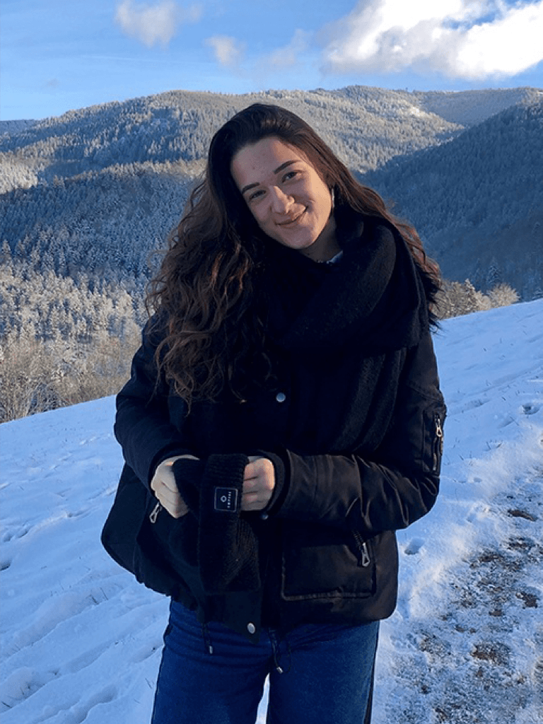 Lara Molz mit langen, braunen Haaren steht in einer schneebedeckten Landschaft. Sie trägt eine schwarze Jacke und einen Schal und lächelt sanft in die Kamera.