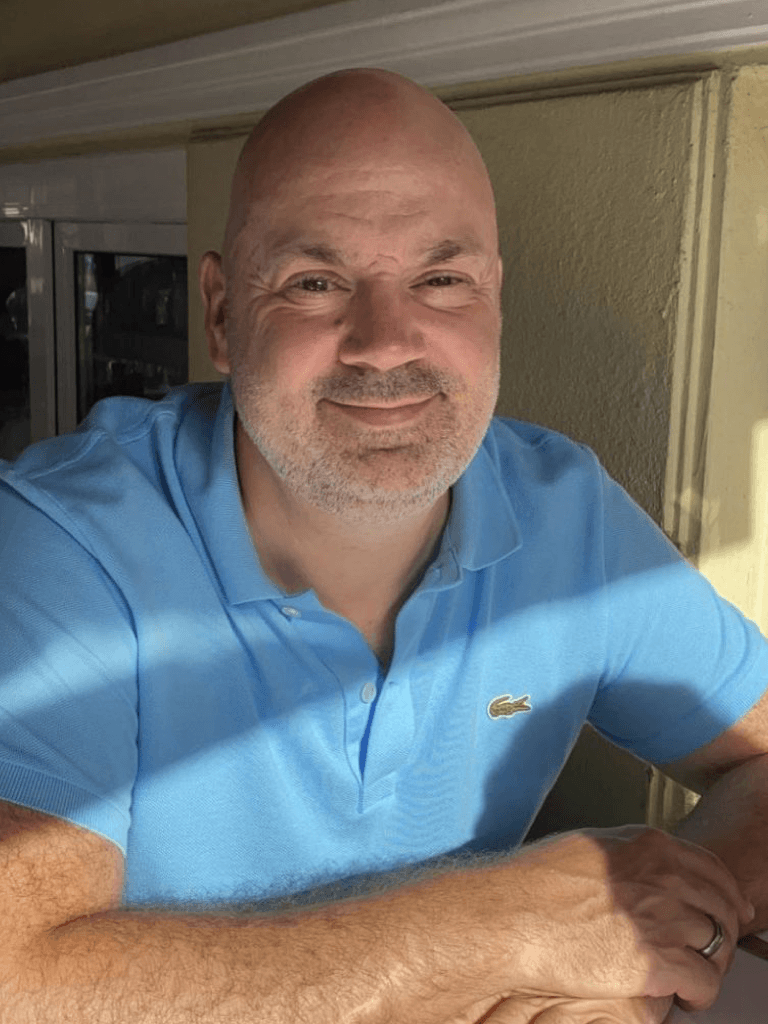 Marko Reiss in einem hellblauen Polohemd sitzt an einem sonnigen Tisch und lächelt in die Kamera. Sonnenlicht fällt auf sein Gesicht und die Umgebung.