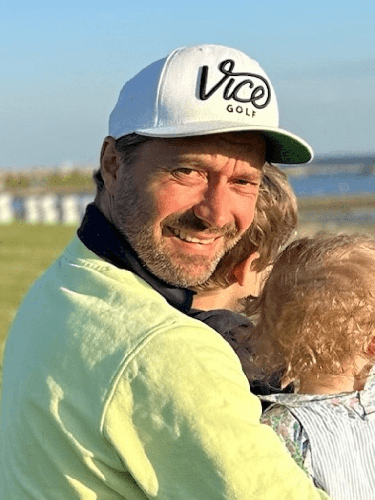 Norman Habenicht mit weißer Baseballmütze hält ein kleines Kind im Arm. Er trägt ein hellgelbes Oberteil und lächelt in die Kamera, während im Hintergrund eine grüne Landschaft und das Meer zu sehen sind.