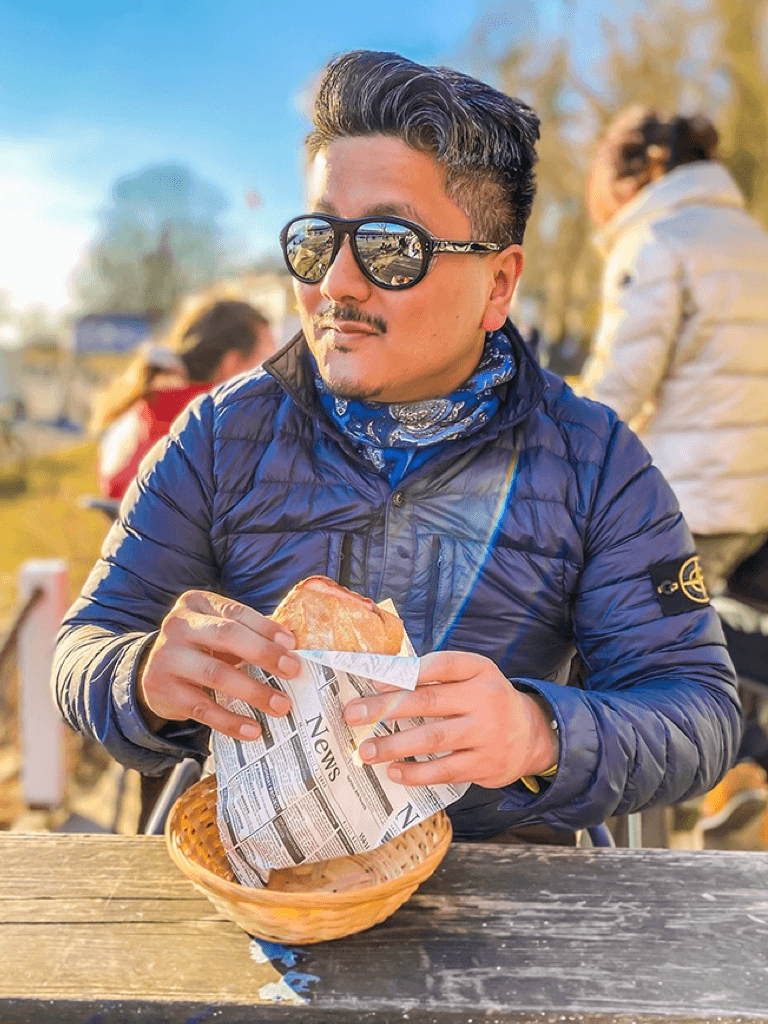 Prasanga Hayakawa mit Sonnenbrille und blauer Steppjacke sitzt an einem Holztisch im Freien und hält ein in Zeitungspapier gewickeltes Sandwich. Im Hintergrund sind andere Personen und eine sonnige Parklandschaft zu sehen.