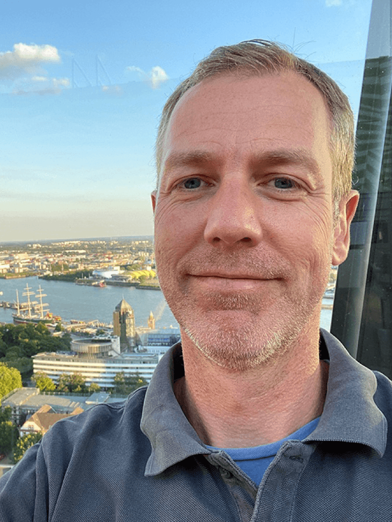 Robert Scheduikat mit kurzem Haar und einem grauen Poloshirt steht auf einer Aussichtsplattform mit Blick auf einen Fluss und die Stadt. Im Hintergrund sind Gebäude, Wasser und ein Schiff zu sehen.