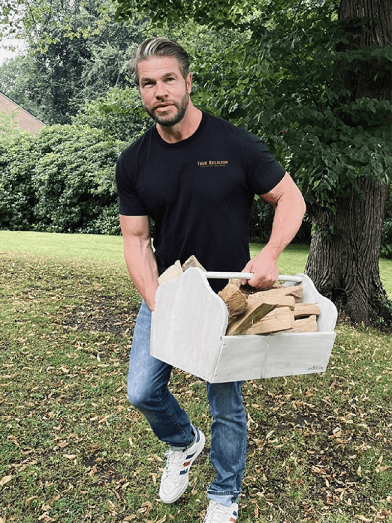 Roland Potthoff in einem schwarzen T-Shirt und Jeans trägt eine weiße Holzkiste voller Brennholz über eine grüne Wiese. Im Hintergrund sind Bäume und ein Backsteingebäude zu sehen.