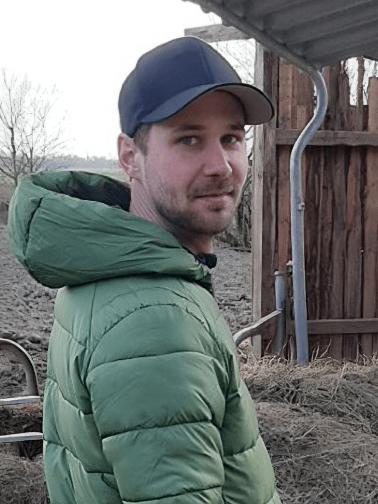 Sebastian Hottenrott mit grünem Steppmantel und dunkelblauer Kappe steht auf einem Hof und blickt über die Schulter in die Kamera. Im Hintergrund sind eine Holzstruktur und ein offenes Feld zu sehen.