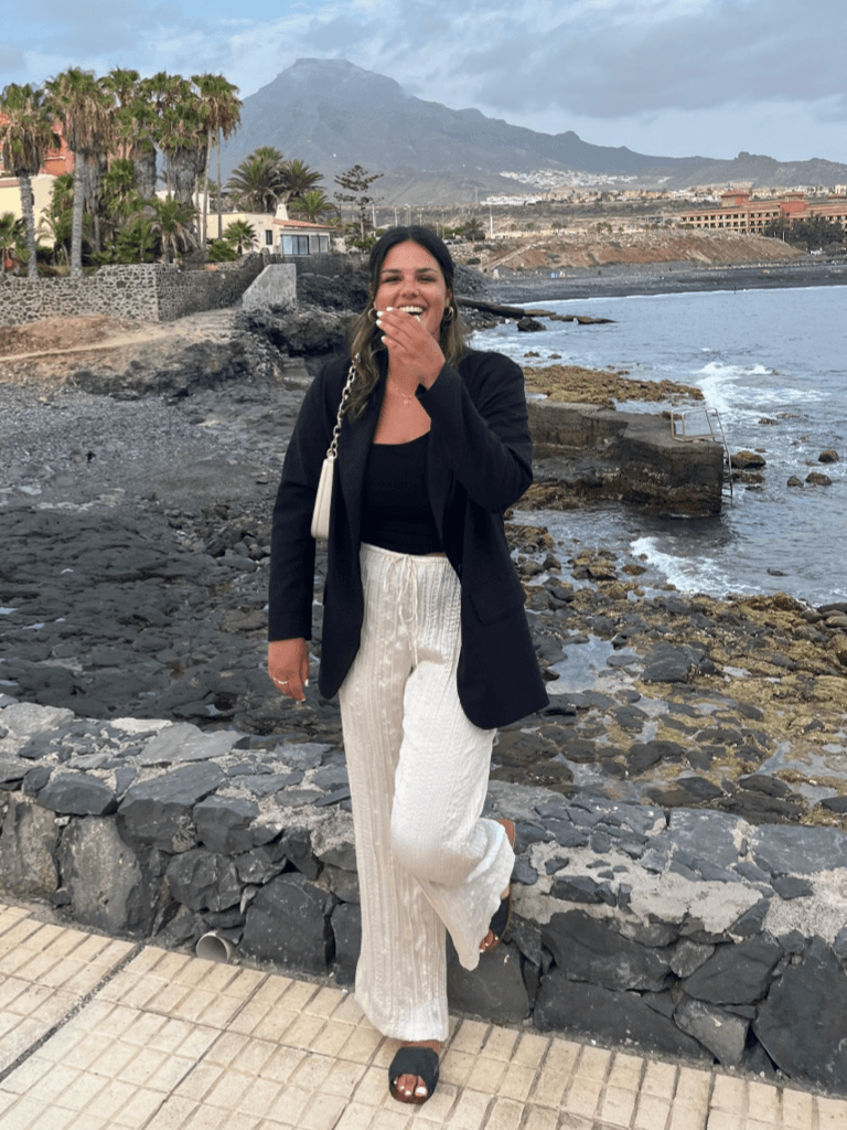 Tatiana Rodriguez in einem schwarzen Blazer und weißen, lockeren Hosen steht lachend an einer Promenade mit Blick auf eine felsige Küste. Im Hintergrund sind Palmen, Gebäude und ein Berg zu sehen, der von Wolken umgeben ist.