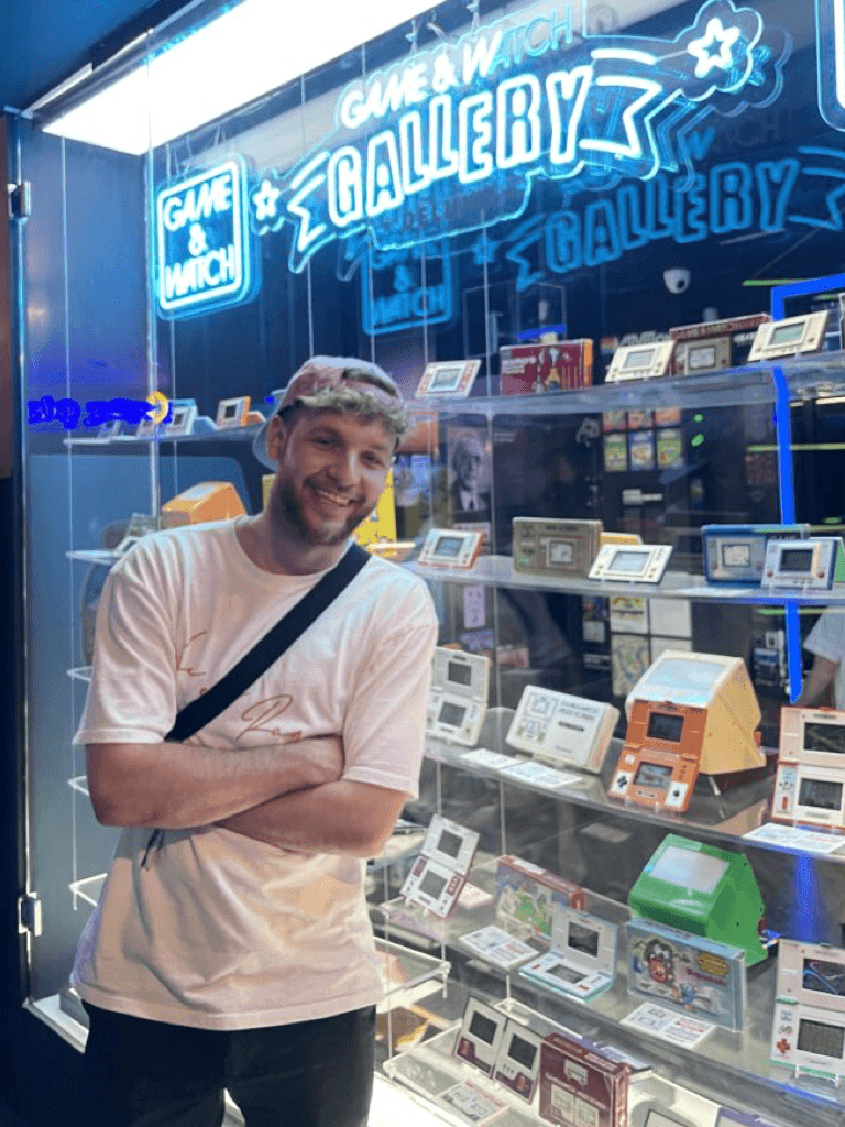 Tim Schippmann mit blondem Haar steht lächelnd vor einer beleuchteten Vitrine mit Retro-Videospielkonsolen und Sammlerstücken. Über der Vitrine leuchtet ein blaues Neon-Schild mit der Aufschrift 'Game & Watch Gallery'
