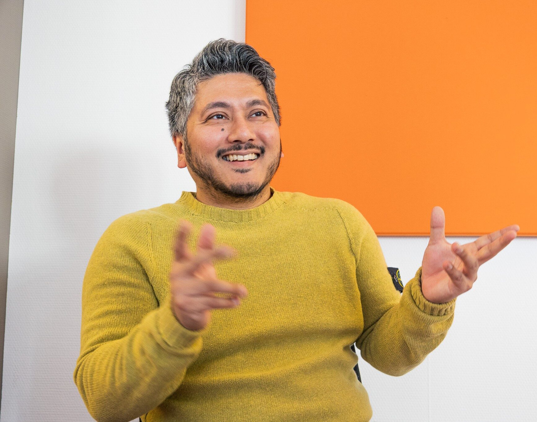 Ein Mann im senfgelben Pullover sitzt vor einer weißen Wand mit orangem Wandpanel. Er gestikuliert lebhaft und lacht dabei – die Szene wirkt kommunikativ und offen.