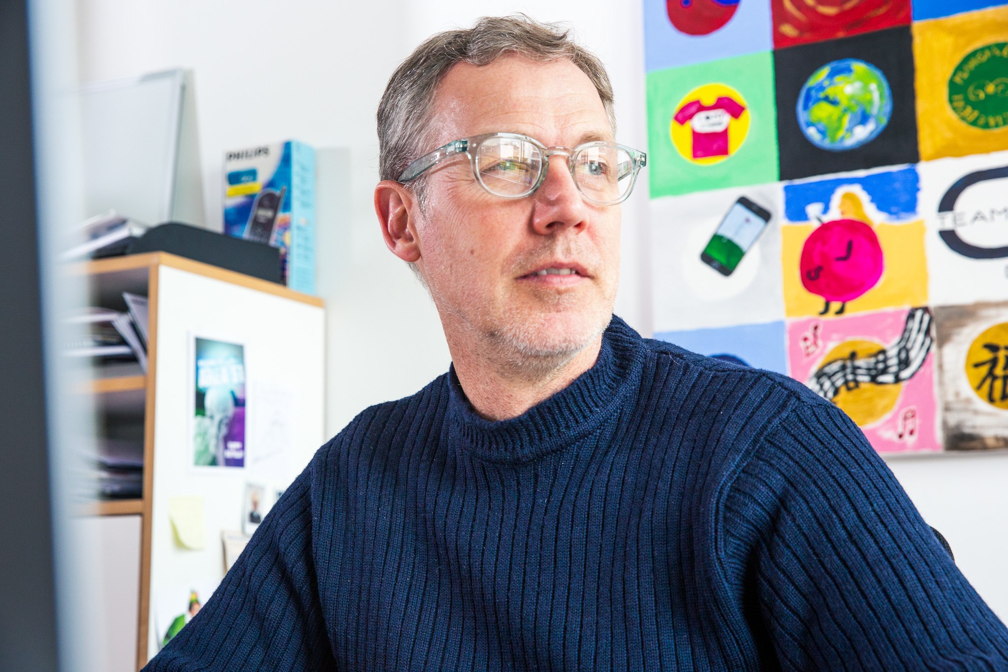 Ein Mann mit Brille sitzt an seinem Schreibtisch und blickt leicht zur Seite. Im Hintergrund sind bunte Illustrationen an der Wand und typische Büromaterialien zu sehen.