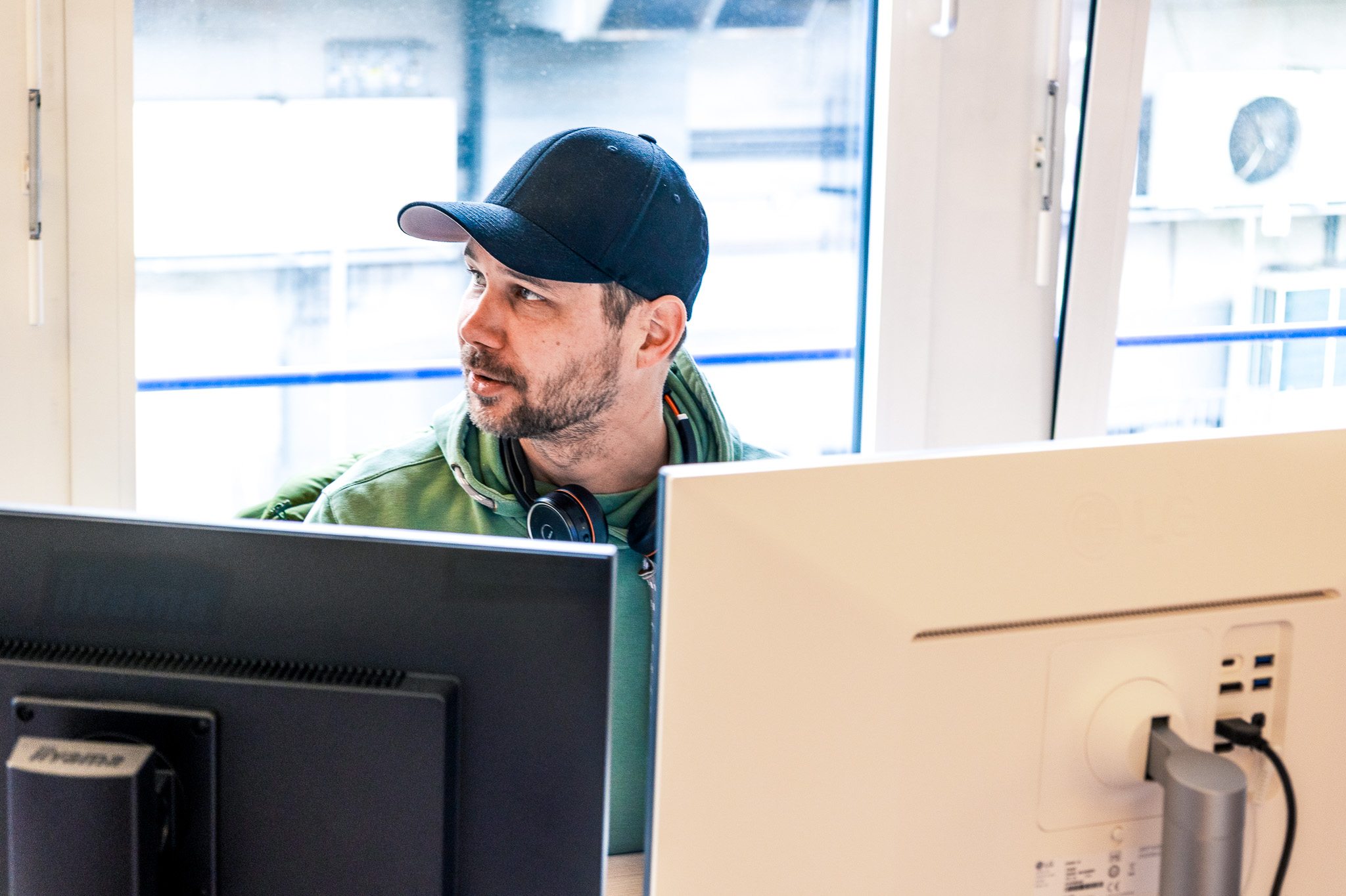 Ein Mann mit dunkler Kappe und Kopfhörern sitzt an zwei Monitoren und blickt zur Seite. Durch das Fenster fällt Tageslicht ins Büro.