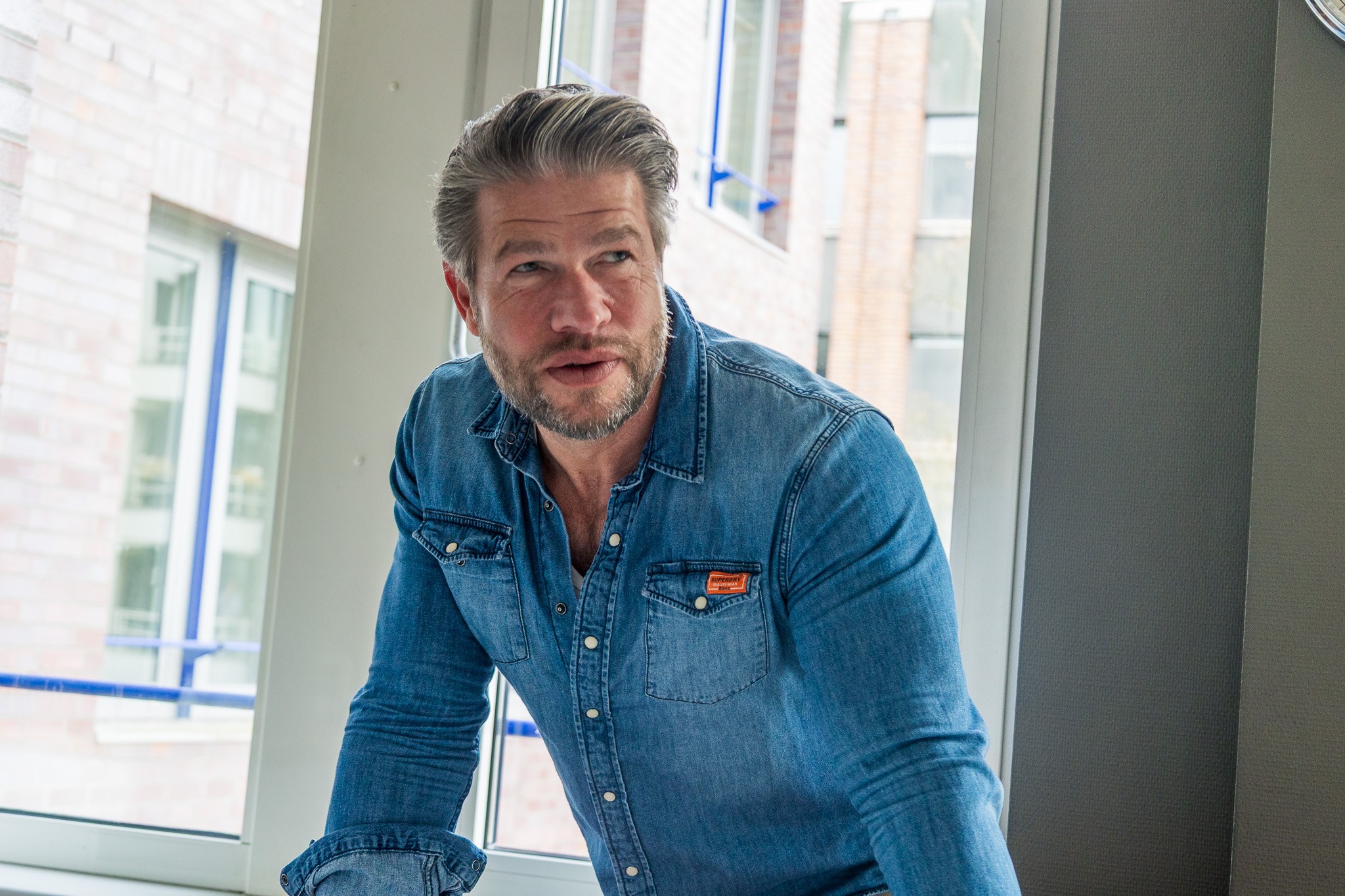Der Mann im blauen Jeanshemd lehnt sich leicht nach vorne und wirkt, als sei er mitten in einem Gespräch. Das Bild wurde vor einem großen Fenster aufgenommen, Tageslicht fällt in den Raum.