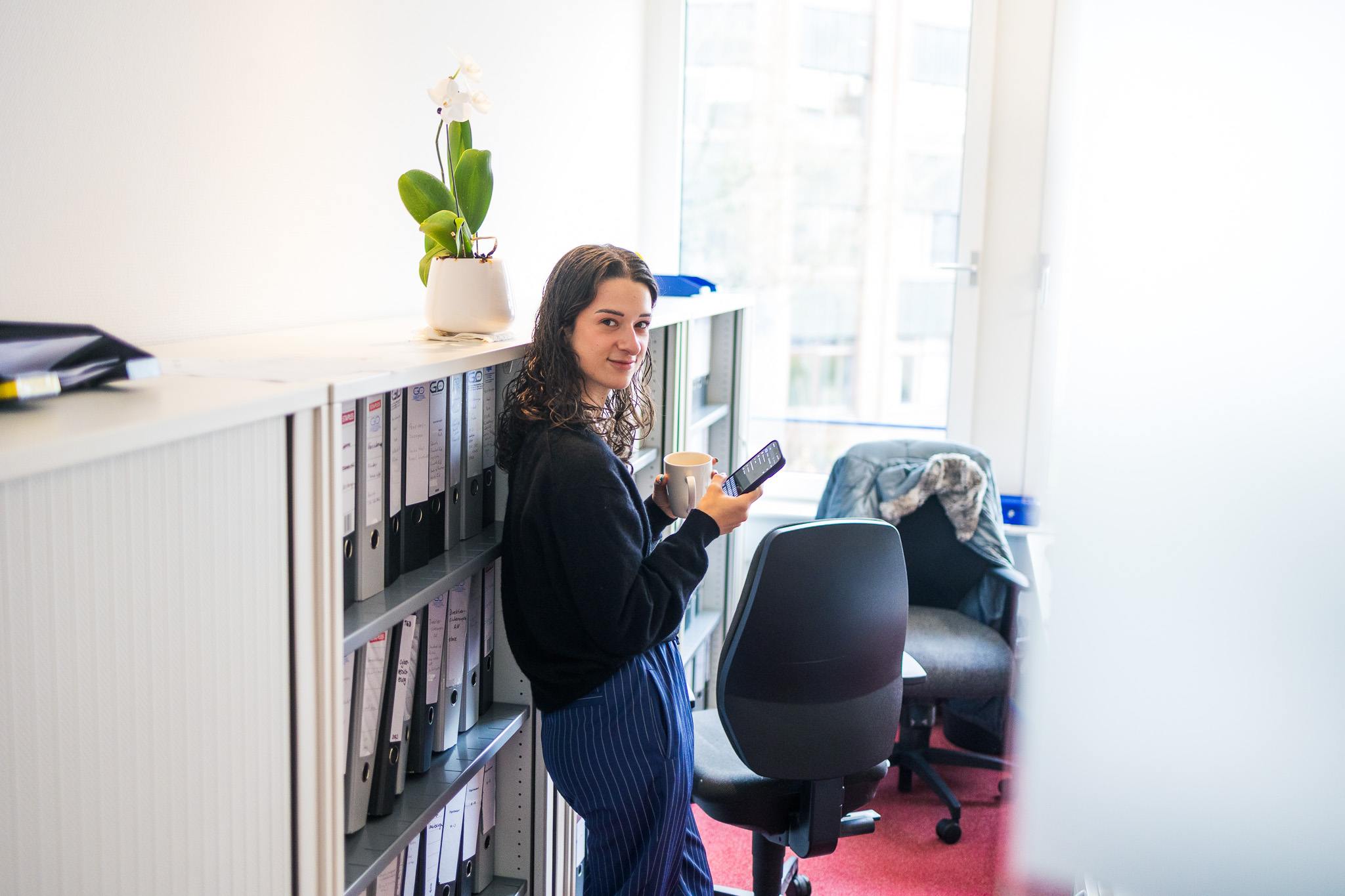 Eine junge Frau steht mit einer Kaffeetasse und einem Smartphone vor Aktenschränken. Sie lächelt in Richtung Kamera. Eine weiße Orchidee steht auf dem Regal, im Hintergrund ein großes Fenster.