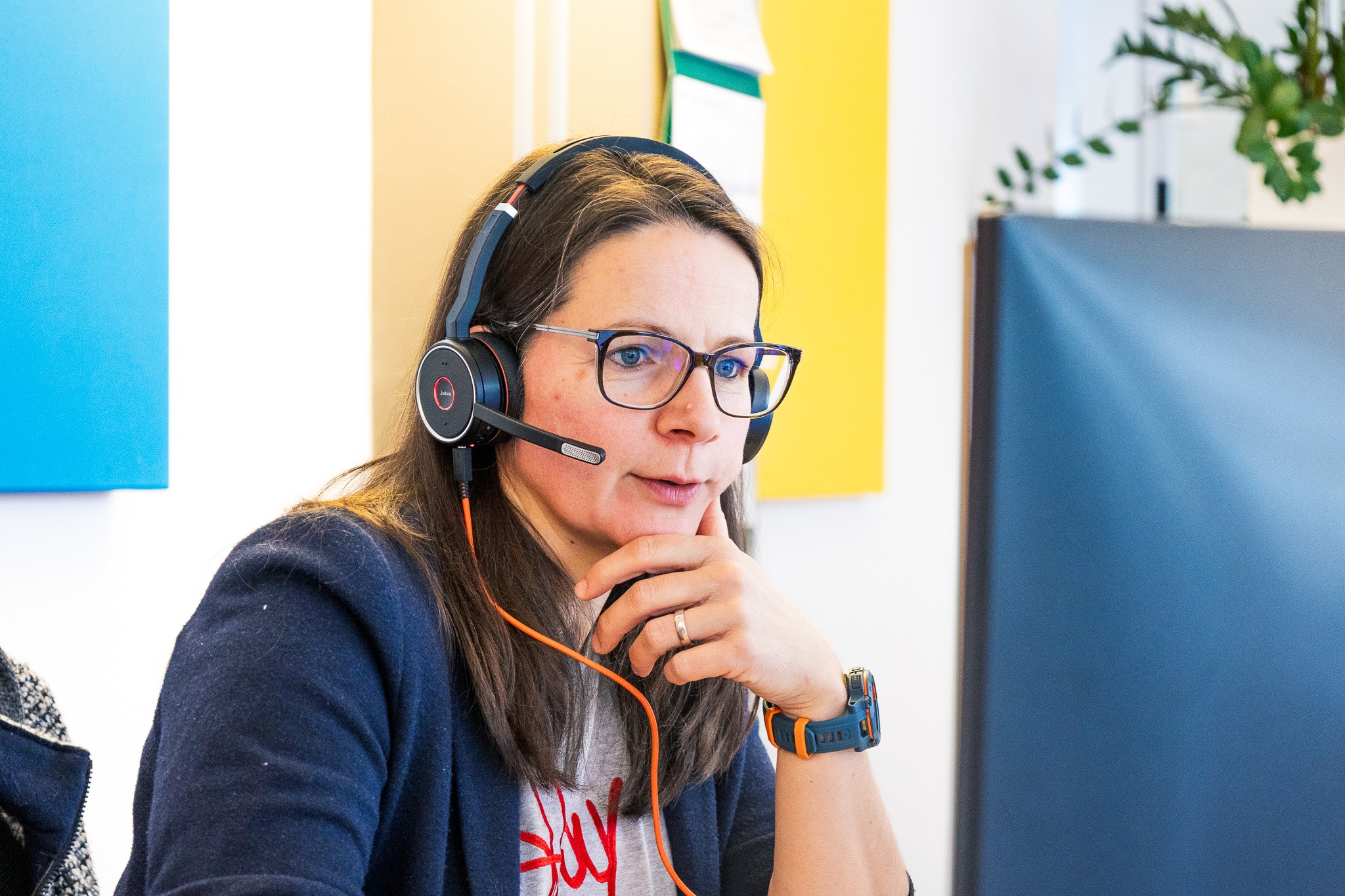 Eine Frau mit Headset und Brille sitzt an einem Schreibtisch und schaut konzentriert auf einen Bildschirm.
