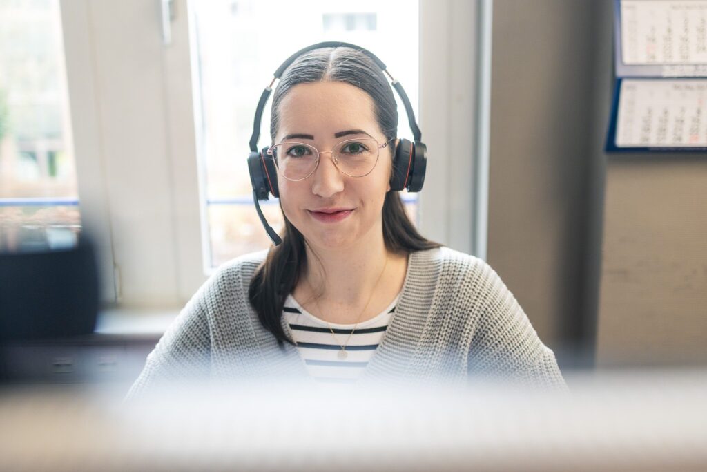Eine Frau mit Headset sitzt an ihrem Arbeitsplatz und schaut direkt in die Kamera. Sie trägt eine Brille und einen hellen Strickpulli. Im Hintergrund ist ein Fenster zu sehen.
