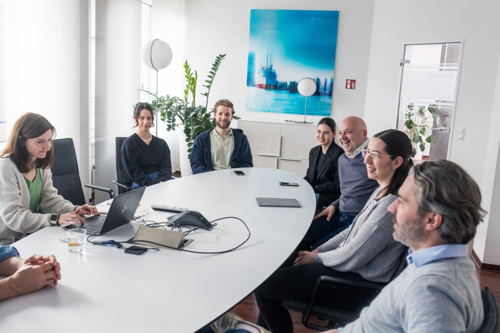 Acht Personen sitzen gemeinsam an einem großen Konferenztisch in einem hellen Raum. Einige lachen, andere hören konzentriert zu – die Atmosphäre ist offen und kollaborativ.