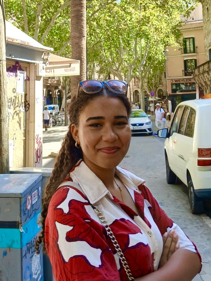 Junge Frau mit geflochtenen Haaren steht lächelnd in einer sonnigen Stadtstraße mit Palmen, Cafés und parkenden Autos im Hintergrund. Sie trägt ein auffälliges, rot-weiß gemustertes Hemd und Sonnenbrillen auf dem Kopf.