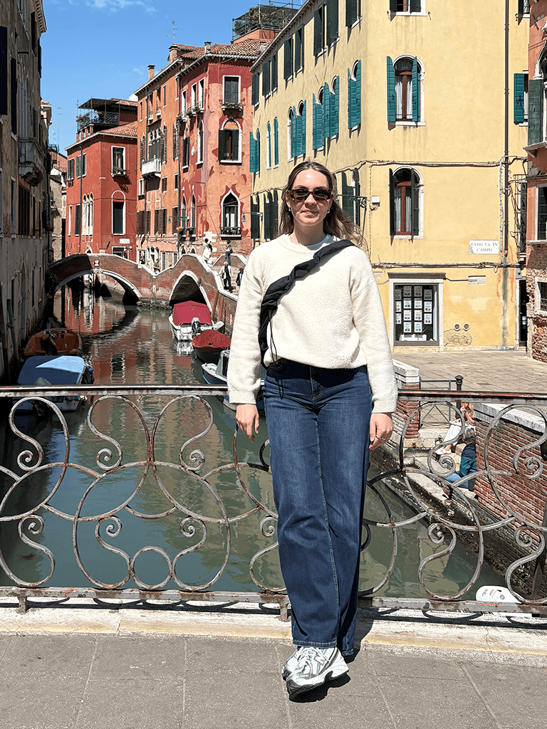 Maren Jansen mit Sonnenbrille und cremefarbenem Pullover steht auf einer venezianischen Brücke vor einem Kanal mit Booten und pastellfarbenen Häuserfassaden.