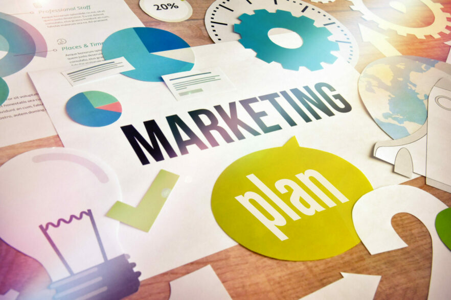 Verschiedene Diagramme und Symbole umgeben das Wort "Marketingplan" auf einem Schreibtisch.